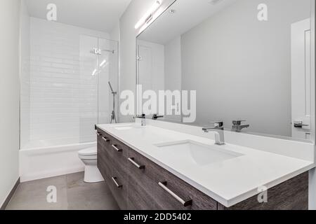 A luxury bathroom with a wood vanity cabinet, white counter top, and chrome faucets with a white tiled shower. Stock Photo