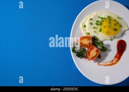 A dish of fried eggs. Blue background with space for text. Fried eggs on a white plate with tomatoes, herbs, ketchup. Bachelor's Breakfast Stock Photo