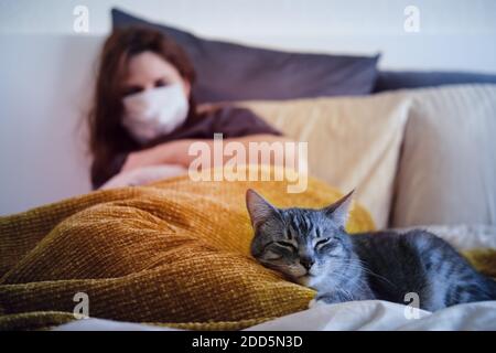 A woman in a medical mask and a cat lie on the bed Stock Photo