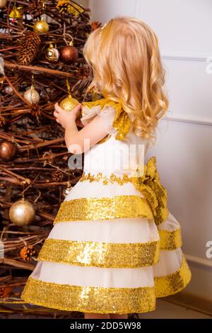 A little girl in a chic white and gold dress stands with her back to the camera and decorates the Christmas tree Stock Photo