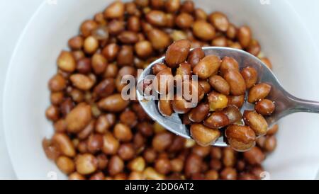 A spoonful of fried peanuts, coated with skin. With a bowl of peanuts in the blurred background. Stock Photo