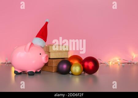 Pink piggy bank in the hat of Santa Claus, gift boxes, Christmas baubles and shining garland on pink background. Savings for Christmas and New Year, h Stock Photo
