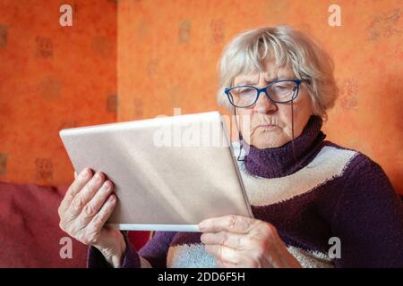 Senior woman in eyeglasses using tablet and sitting on couch at home. Grandmother holding digital device in hands. Gray hair old lady looking at table Stock Photo