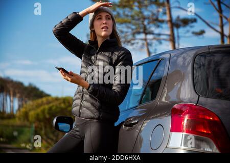 Young woman standing next to car which has broken down or she could be lost and trying to get signal on mobile phone to use map app or call for help Stock Photo