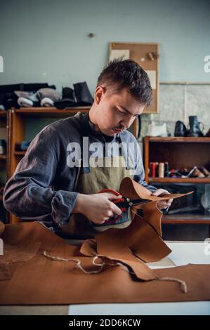 A young tanner in his workshop at the table cuts out leather elements of shoes. Stock Photo