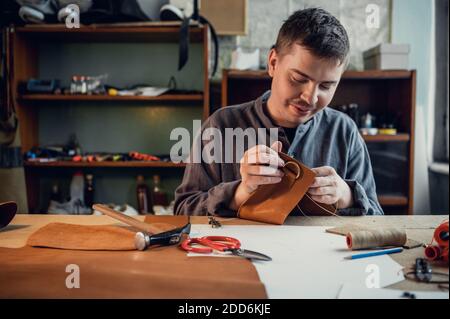 A young shoemaker manually sews decorative elements to leather shoes in the workshop. Stock Photo