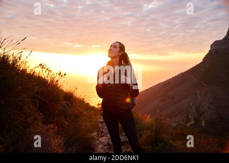 Front view of young woman on vacation wearing backpack setting off on hike climbing coastal path at sunset Stock Photo