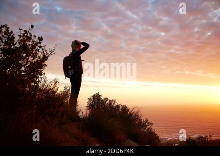 Side view of young woman on vacation wearing backpack hiking along coastal path at sunset looking out to sea Stock Photo