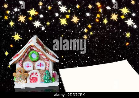 Lamp in the form of a gingerbread house and a blank sheet of paper for text, on a black background with falling snow and superimposed stars Stock Photo