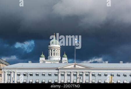 Main green dome of white Helsinki Cathedral and top of Helsinki City Hall with the Flag of Finland in the roof against dark stormy sky.