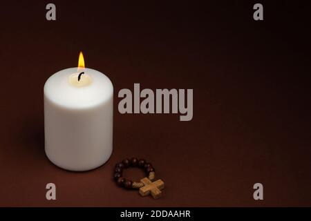 One white burning candle and wooden cross on dark background with copy space for text. Stock Photo