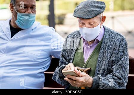 Senior man using smart phone wearing protective face mask sitting with mature male on bench during COVID-19 Stock Photo