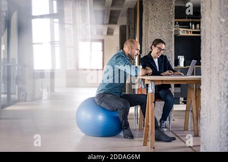 Businesswoman using laptop by man sitting on fitness ball at office