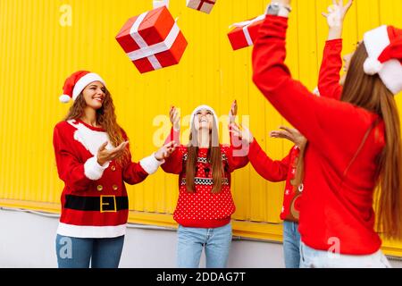 Excited, rejoicing group of friends wearing santa claus hats having fun and tossing christmas presents on yellow wall Stock Photo