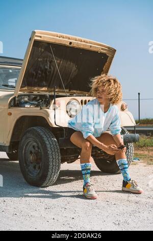 Young blond Afro woman looking away while sitting on bumper of broken-down vehicle during sunny day Stock Photo