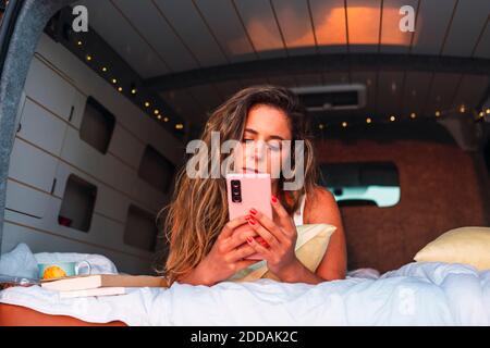 Beautiful woman using mobile phone while lying in camper van during sunset at beach Stock Photo