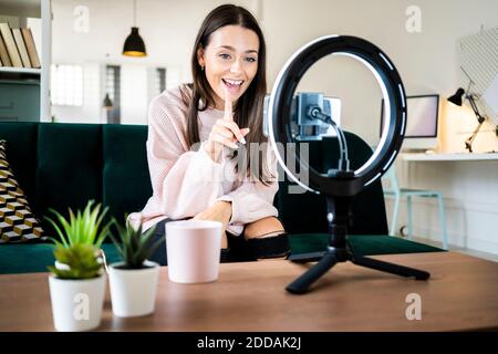 Happy beautiful female vlogger sitting on sofa while filming through smart phone at loft apartment Stock Photo