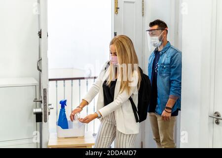 Blond woman wearing face mask washing hands while standing with coworker in background at office