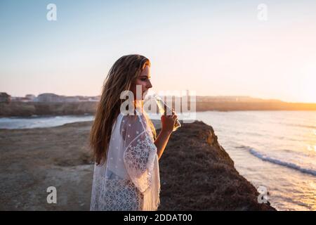 Young woman with small white wine bottle looking away while standing at beach Stock Photo