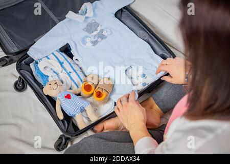 Young pregnant woman packing suitcase and baby clothes at home to go to maternity hospital - Pregnancy, birth concept Stock Photo