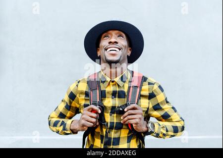 Smiling african man with backpack in hat looking up against white wall Stock Photo