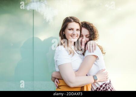 Smiling beautiful woman being hugged by young female friend against wall Stock Photo