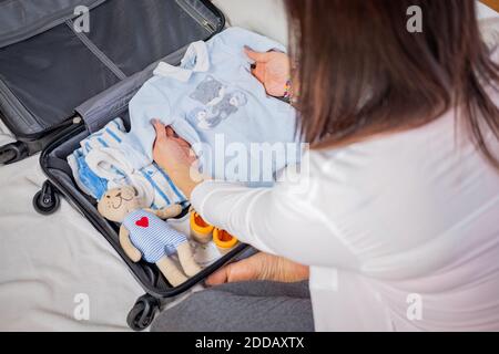 Young pregnant woman packing suitcase and baby clothes at home to go to maternity hospital - Pregnancy, birth concept Stock Photo