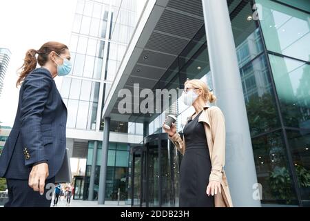 Business people wearing face mask talking while standing with social distance against office building Stock Photo