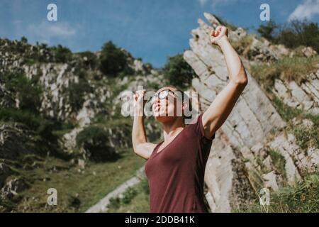 Woman celebrating victory with arms raised against mountain on sunny day Stock Photo