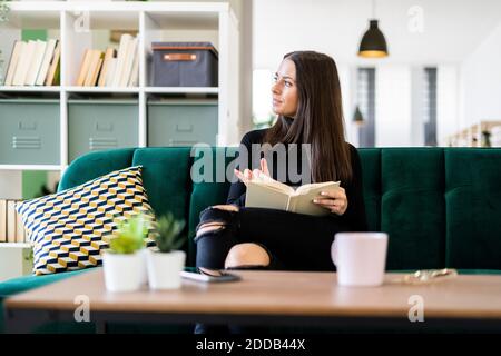 Thoughtful young woman looking away while sitting with book on sofa at home Stock Photo