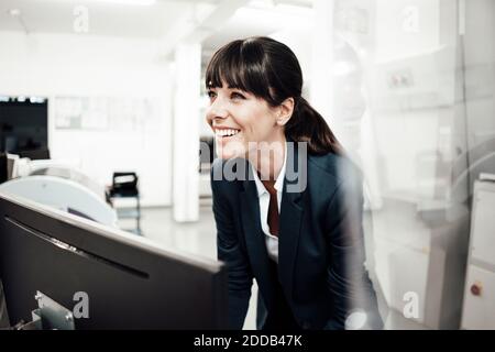 Cheerful businesswoman looking away while standing in front of computer monitor at industry Stock Photo