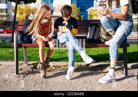 Low section of mother with kids using smart phone while sitting on bench in public park during sunny day Stock Photo