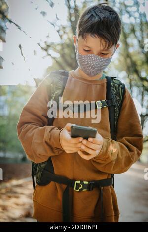 Boy using smart phone wearing protective face mask while standing in public park Stock Photo