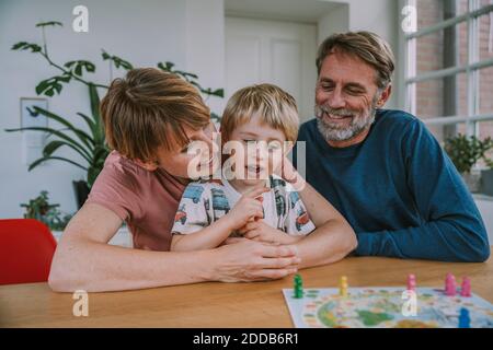 Mother embracing son while father sitting beside playing board game at home Stock Photo