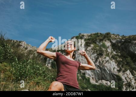 Smiling woman celebrating victory with arms raised against sky on sunny day Stock Photo