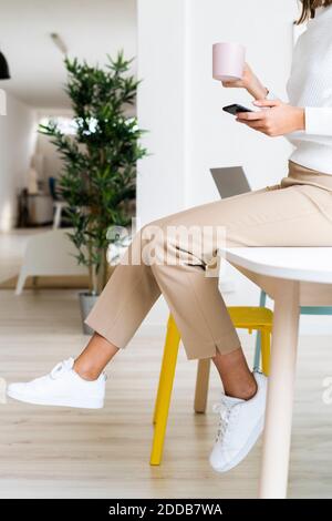 Businesswoman with coffee cup using mobile phone while sitting on table at office Stock Photo