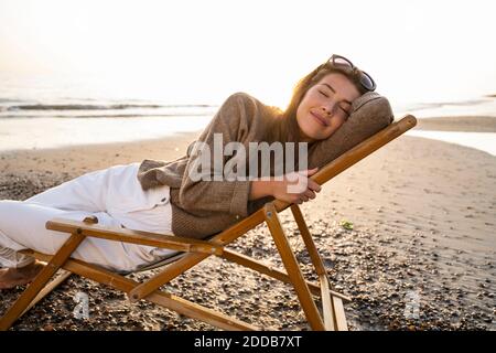 Smiling beautiful young woman reclining on folding chair while relaxing at beach during sunset Stock Photo