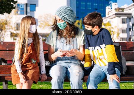 Mother and kids wearing protective face mask using smart phone sitting on bench in public park during sunny day Stock Photo