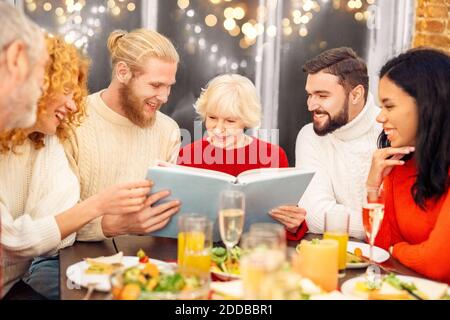 United mixed race family spending time together, looking photo album, laughing and sitting behind served dinner table with homemade food Stock Photo