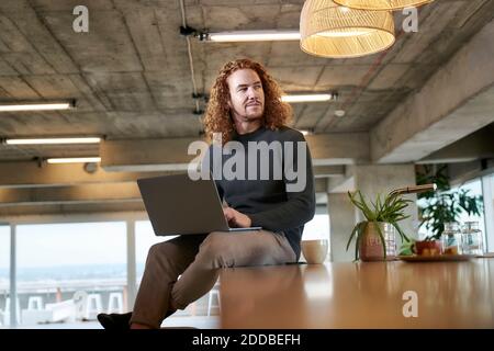 Thoughtful redhead man looking away while using laptop sitting at home Stock Photo