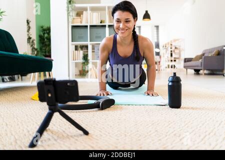 Smiling young woman video recording on camera while doing push ups at home Stock Photo