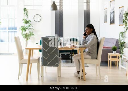 Young woman talking on smart phone while studying through laptop at home Stock Photo