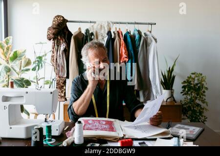Smiling male tailor talking through smart phone while looking at design on paper Stock Photo