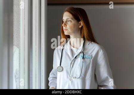 Thoughtful young female doctor looking through window while standing at home office Stock Photo