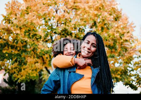 Smiling mother piggybacking daughter while standing at park Stock Photo