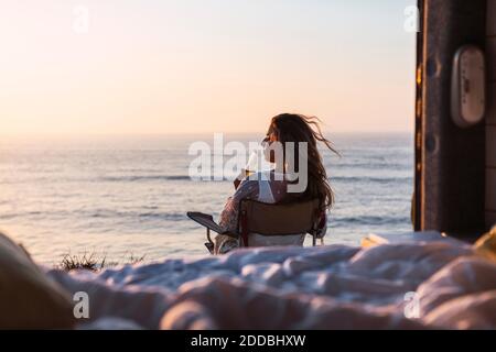 Young woman drinking white wine while sitting on chair by camper van at beach during sunset Stock Photo