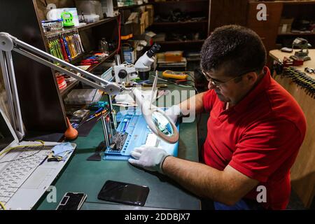 Worker looking at broken smart phone through magnifying glass in repair shop Stock Photo