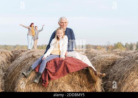 Grandfather and granddaughter sitting on hay bale while grandmother and grandson in background Stock Photo