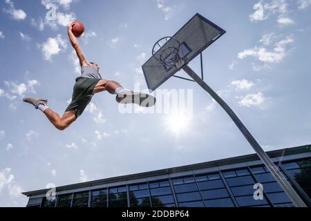 Young man dunking ball in hoop while playing basketball against sky on sunny day