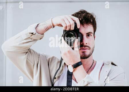Confident man taking photograph through camera while standing against wall Stock Photo